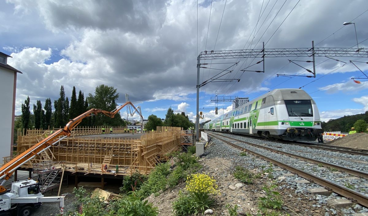 Rakenteilla oleva silta, jossa on puinen tukirakennelma. Sillan vieressä kulkee kaksikerroksinen juna, joka liikkuu sähköradalla. Taustalla näkyy pilvinen taivas ja puita.