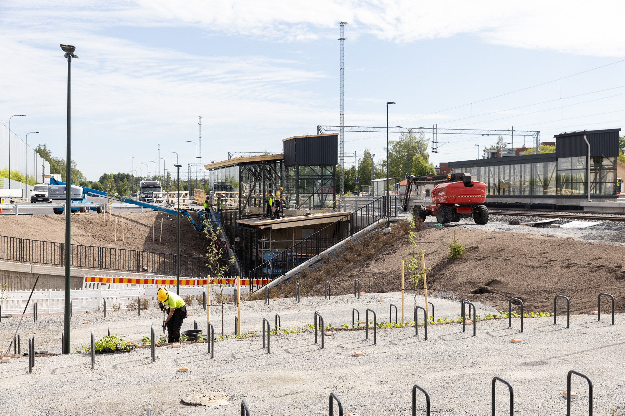 Kuvassa Nokian asema-aluetta kesäisessä maisemassa. Etua-alalla uudistuneet pyöräilytelineet ja henkilö tekemässä vihertöitä. Oikealla rata-aluetta työkoneineen. 