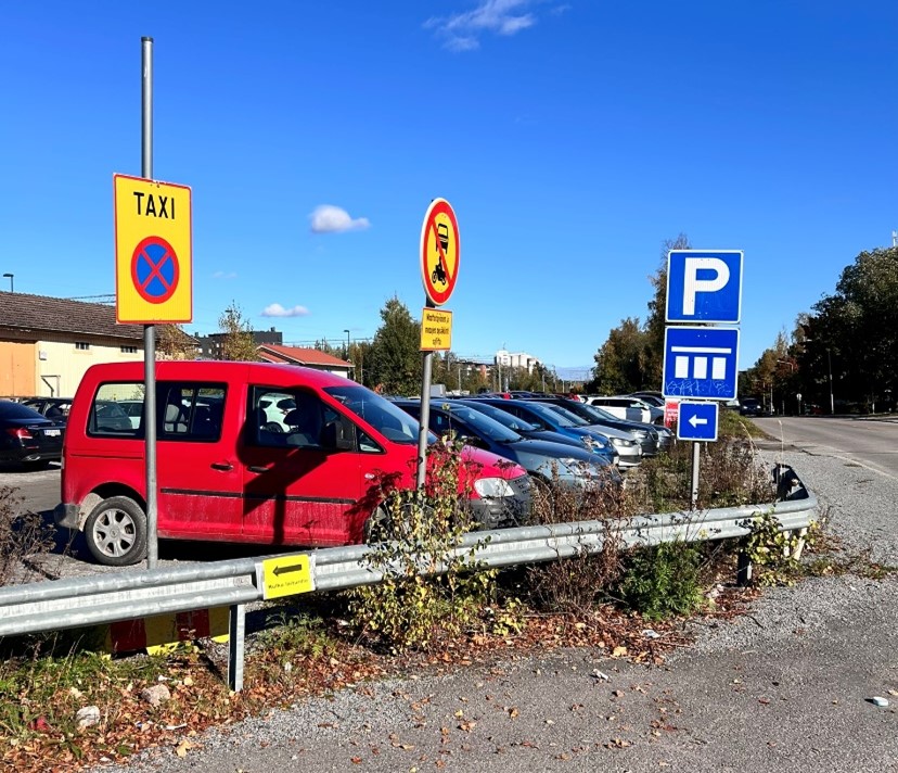 Nokian ratapihan pysäköintialue aurinkoisessa säässä. Etualalla liikennemerkkejä ja autoja. 