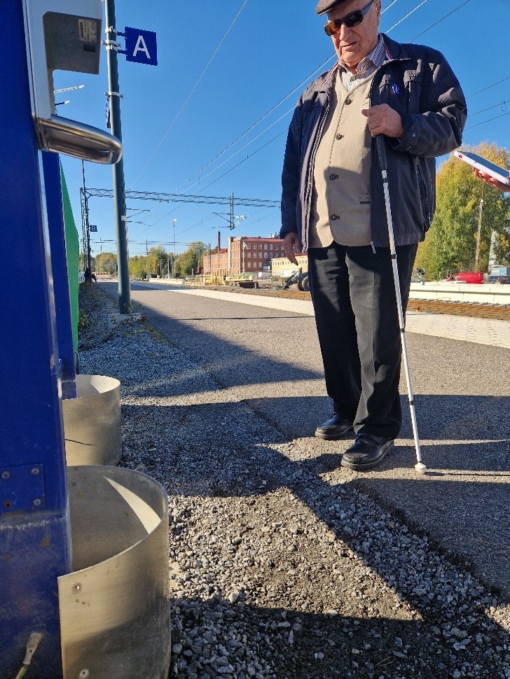 Mies seisomassa laiturialueella kepin kanssa. Oikealla puolella lippuautomaatti ja taka-alalla rataa. 
