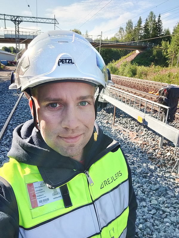Työmaalla oleva henkilö, jolla on päässään valkoinen suojakypärä ja yllään keltainen huomioliivi. Taustalla näkyy rautatie sekä rautatiesilta.