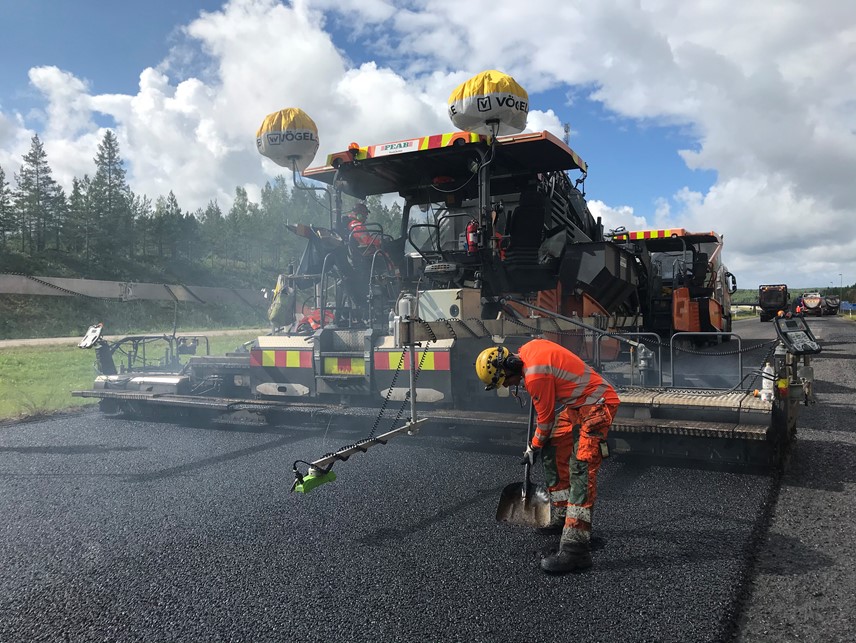 Maanteiden päällystystyöt ovat meneillään. Työntekijä työskentelee lapion kanssa uuden asfaltin päällä. Työkone on taustalla. 