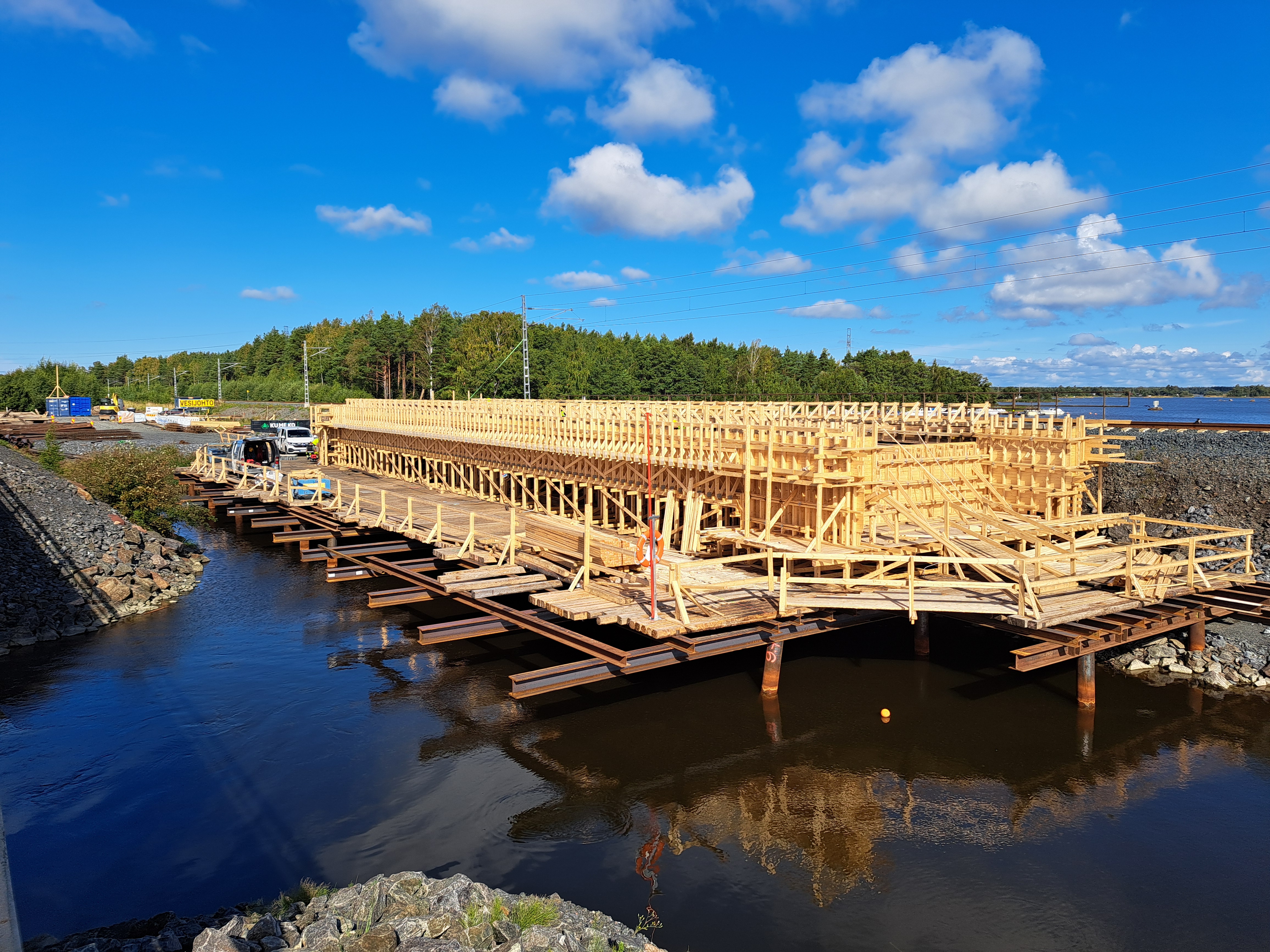 Veden yläpuolella puusta rakennettu sillan muotti, taustalla junarataa ja puita