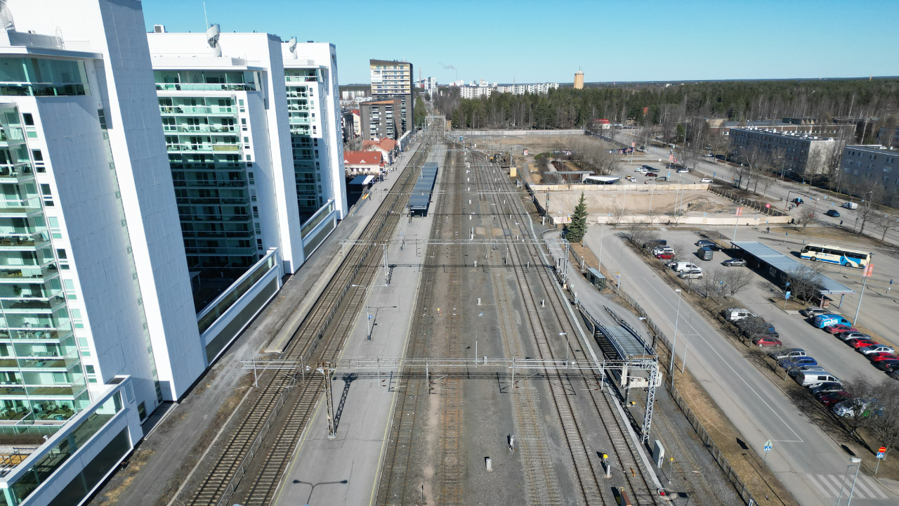 Ilmakuva Oulun rautatieasemasta. Useita rinnakkaisia junaraiteita kulkee pitkittäin kuvan keskellä. Vasemmalla on kolme korkeaa kerrostaloa lasiparvekkeineen. Oikealla tie, pysäköityjä autoja ja bussipysäkki. Taustalla näkyy metsää ja matalampia rakennuksia kirkkaan sinisen taivaan alla.