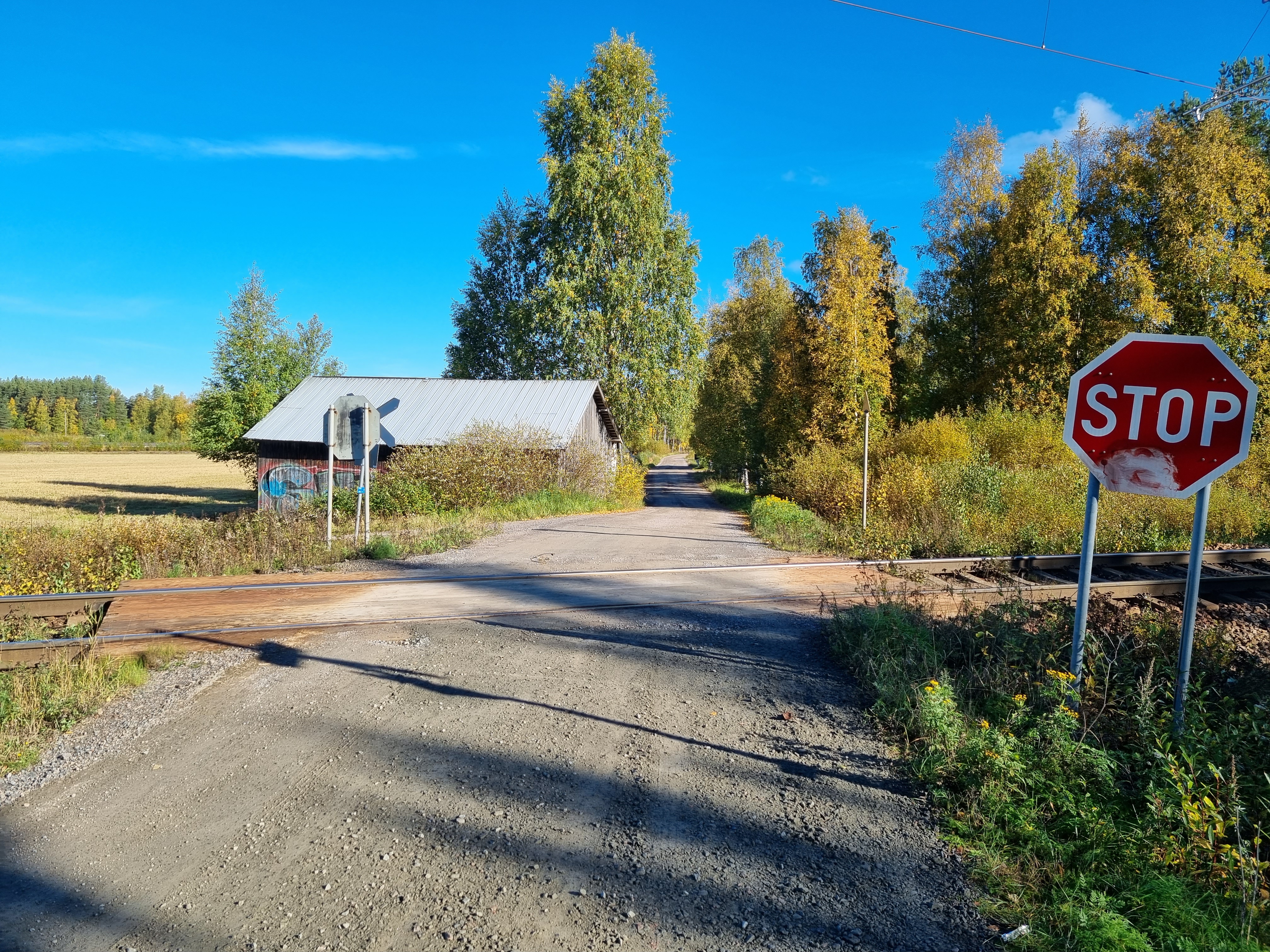 Sorapintaiselta tieltä otettu kuva tasoristeyksestä, jossa tie risteää junaradan kanssa. Radan edessä on STOP-merkki. Radan takana näkyy taivas, peltomaisemaa, lato ja puita.