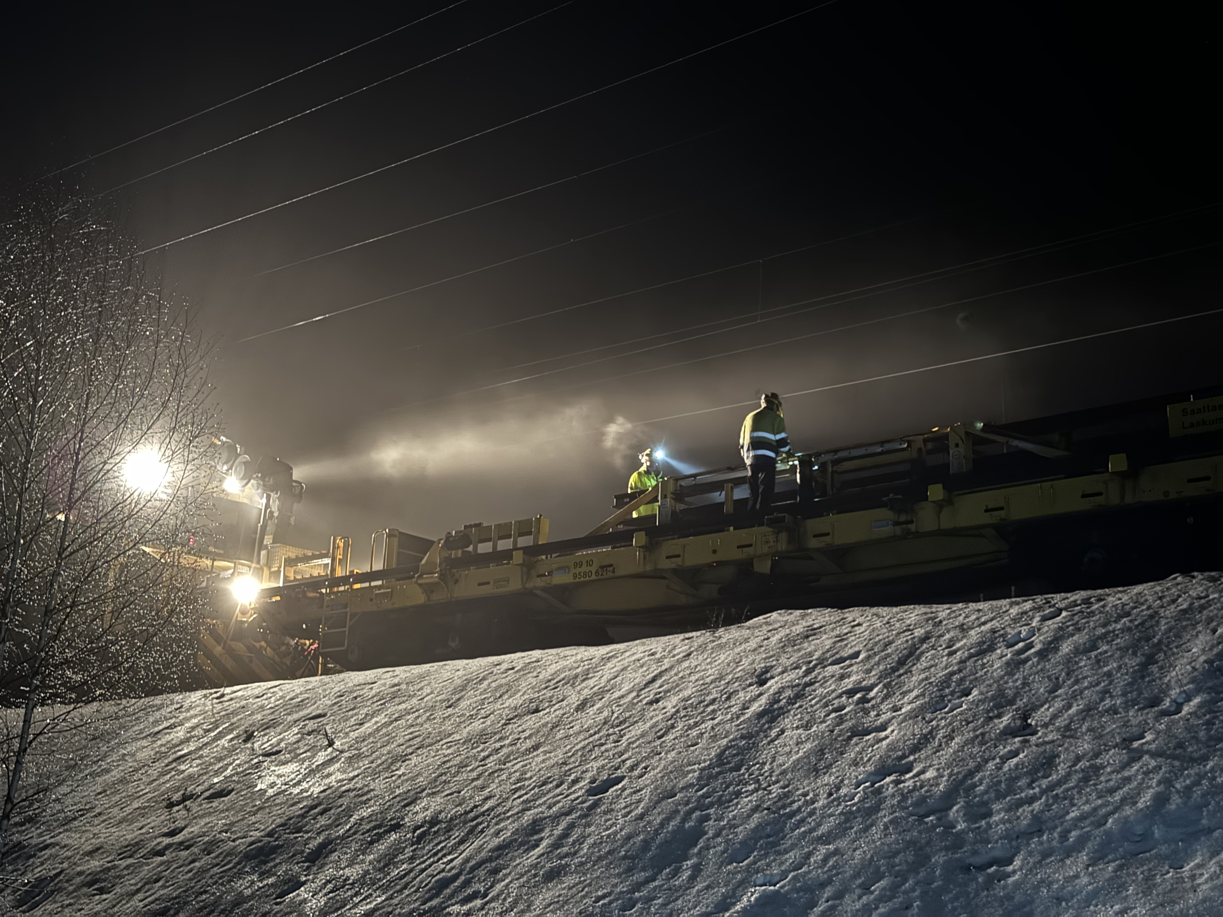Kaksi henkilöä työskentelee talvella pimeässä radalla kiskonjakokoneella