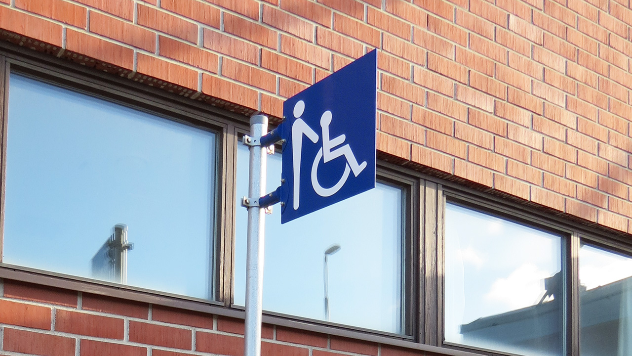 Punatiilisen rakennuksen ulkoseinällä tolppa, jossa sinipohjaisessa merkissä pyörätuolin ja saattajan hahmot.