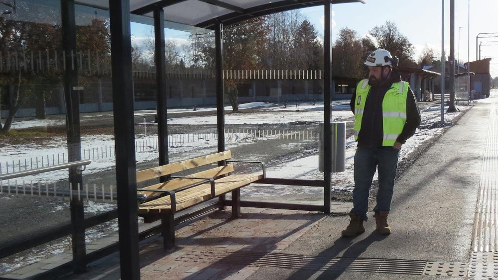 Huomioliiviin pukeutunut työnjohtaja seisoo asemalaiturilla pysäkkikatoksen vieressä, laiturin asfalttipinnassa erottuvat tuntopolut.