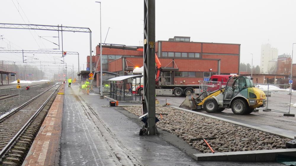 Rakentajat viimeistelevät uutta asfalttipintaa asemalaiturilla, jonka vasemmalla puolella näkyy rata ja oikealla taustalla punatiilinen asemarakennus.