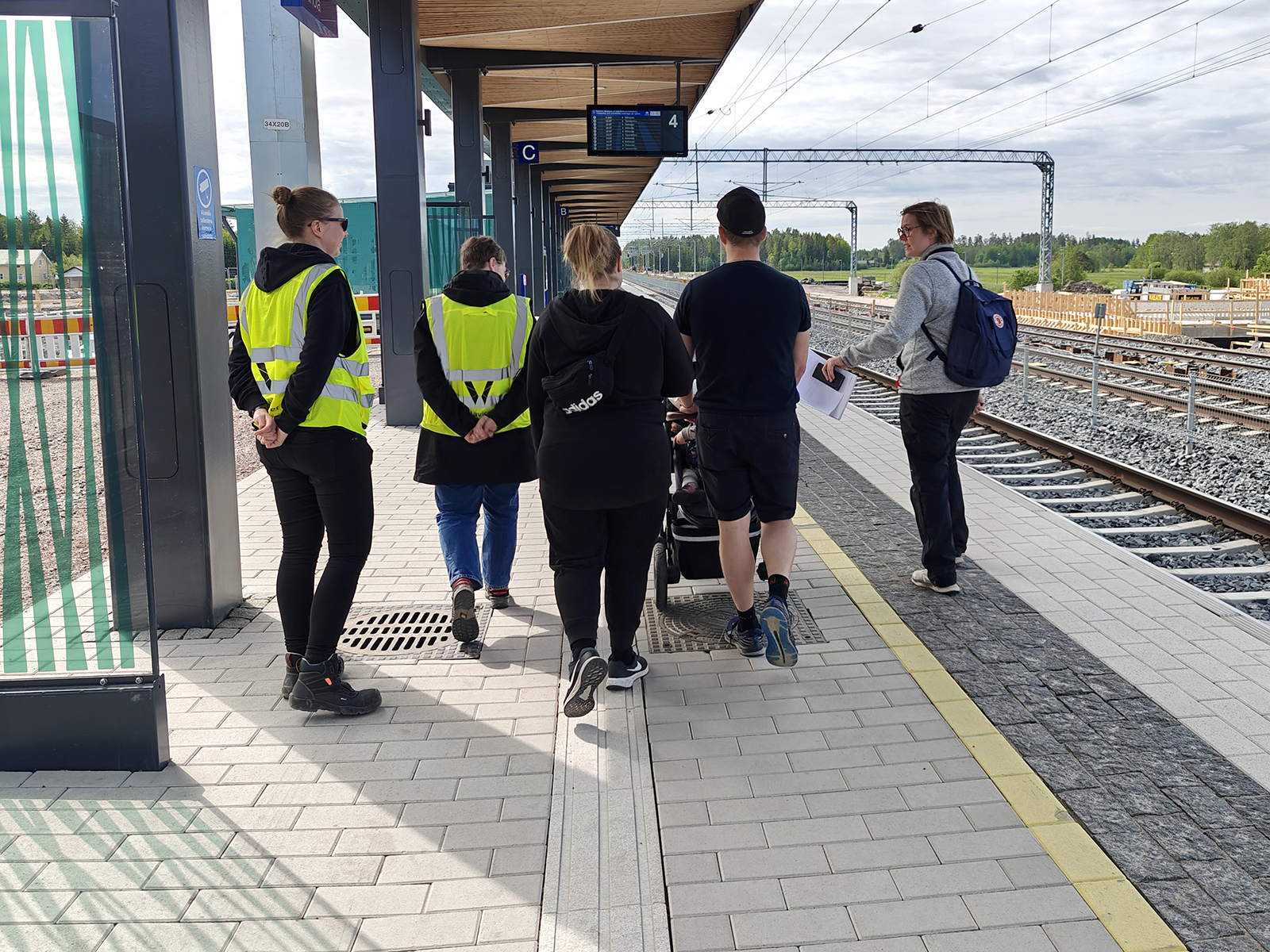 Ryhmä ihmisiä, osa huomioliiveissä, seisoo Ainolan junalaiturilla katsellen kohti raiteita ja keskustellen, taustalla rakennustöitä ja viheralueita.