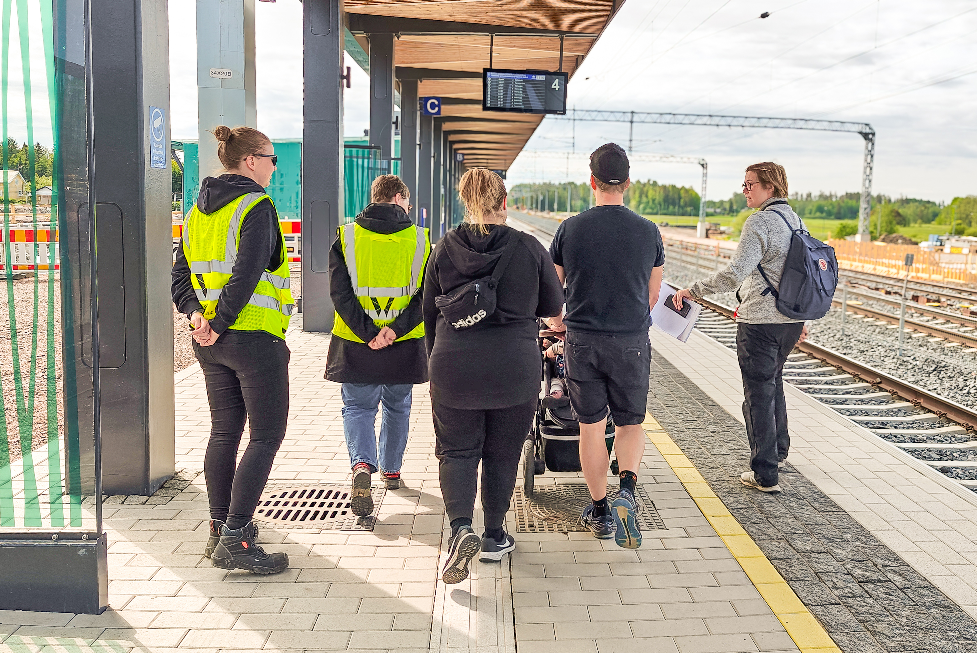 Viisi henkilöä ulkona asemalaiturilla katoksen alla, kahdella huomioliivit, yhdellä lastenrattaat.