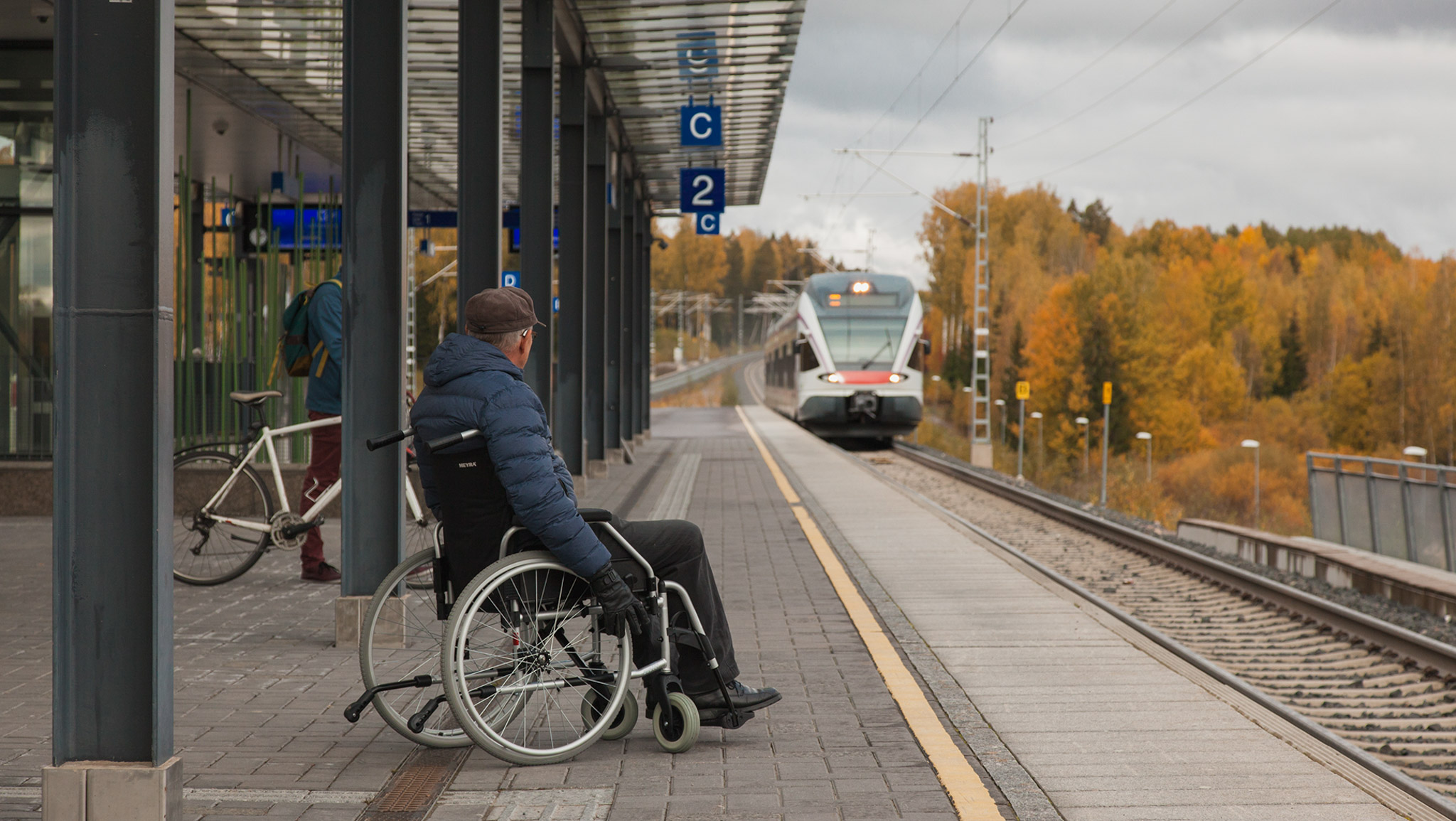 Juna saapuu asemalle syksyllä. Asemalaiturilla mies pyörätuolissa odottaa junaa saapuvaksi.