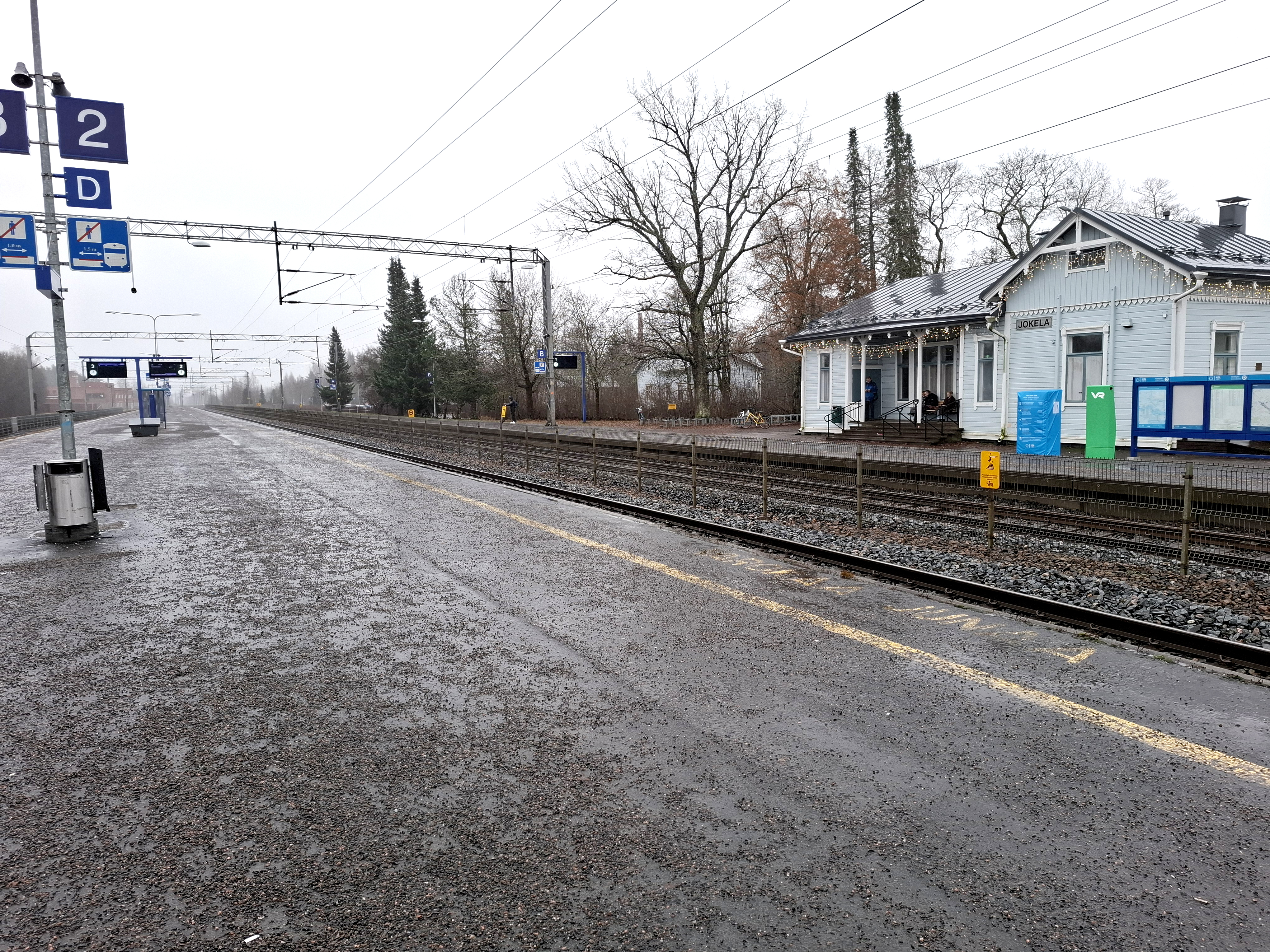 Syksyinen päivä, etualalla junaraiteet ja lähtölaituri 2, taustalla vaaleansinisen sävyisenä Jokelan puinen asemarakennus, jossa kausivalot räystäissä.
