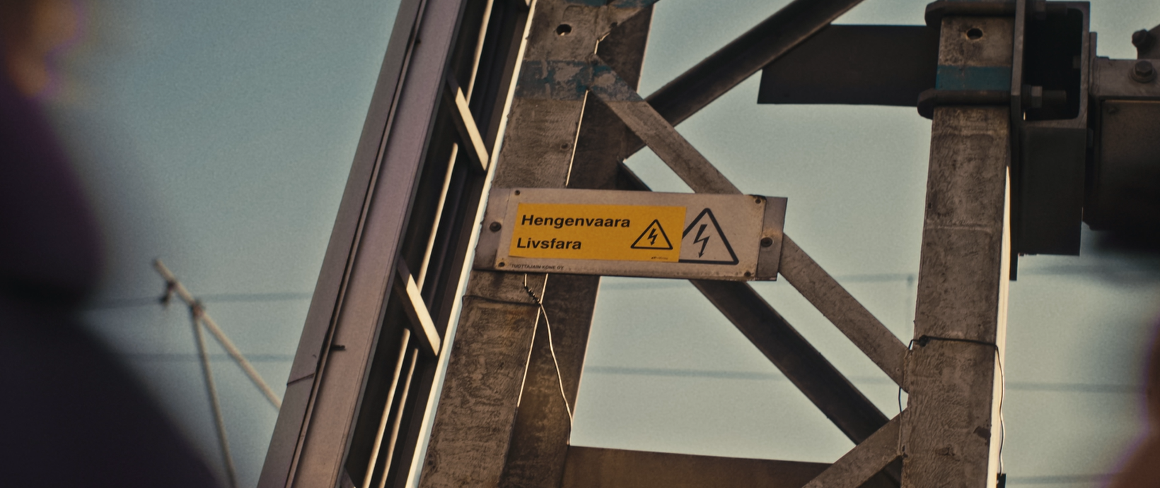 En gul varningsskylt med texten "Livsfara" och en blixtsymbol monterad på en metallkonstruktion. Skylten indikerar fara för elektrisk stöt och högspänning.