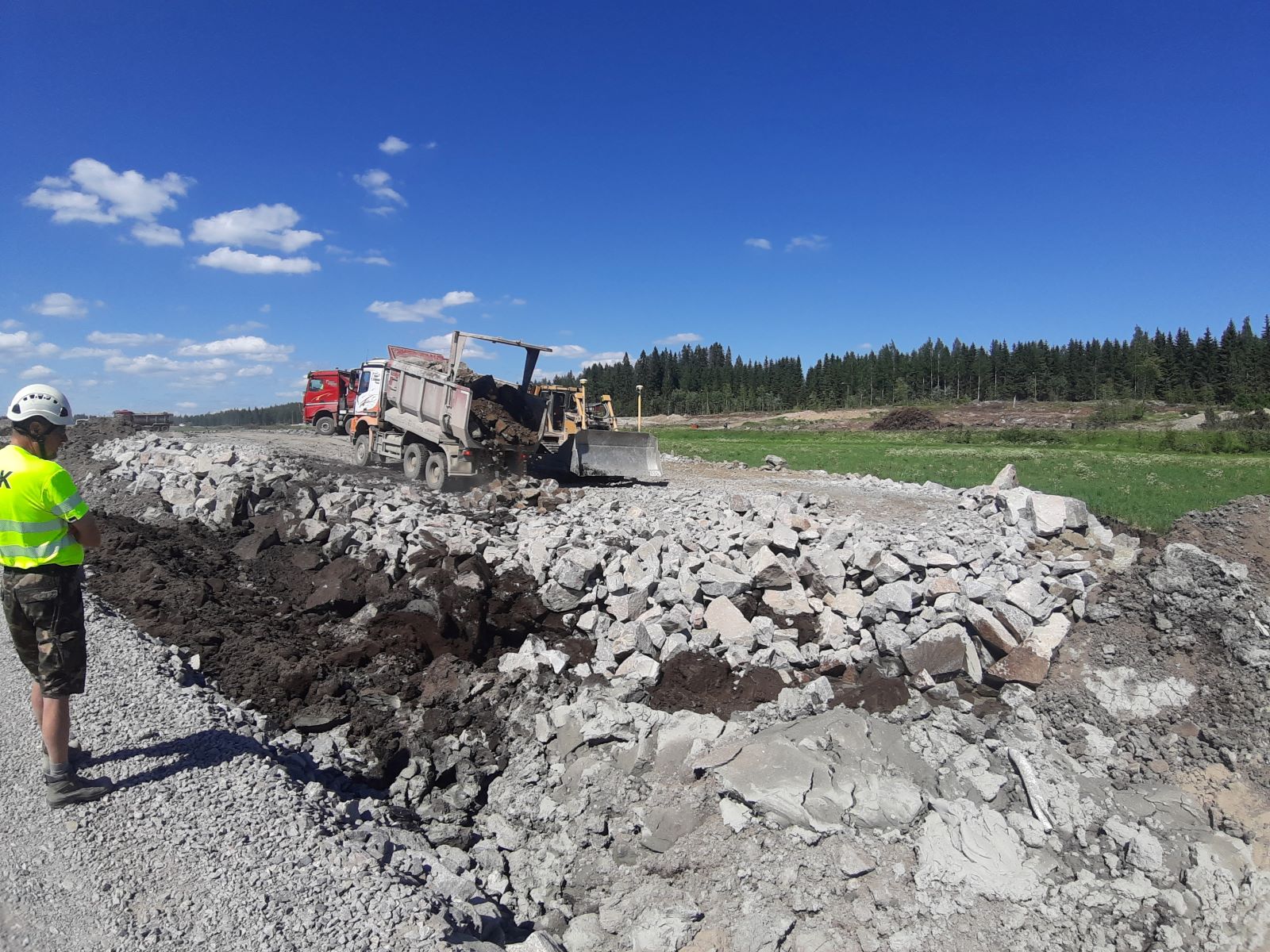 Mies keltaisessa huomioliivissä ja kypärässä katselee maansiirtokoneita, jotka työskentelevät kivisen ja multaisen maan parissa aurinkoisena päivänä metsäisellä alueella.