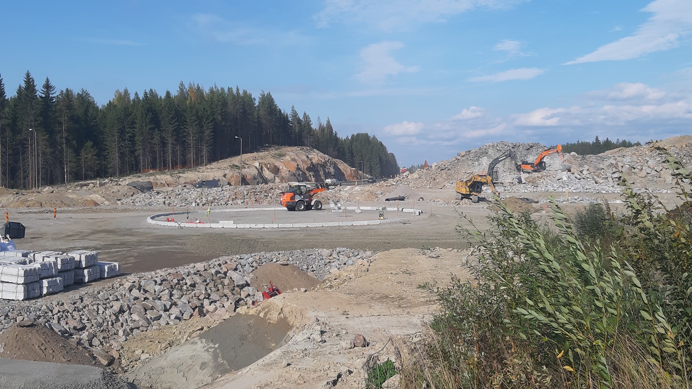 Rakennustyömaa, jossa on keskeneräinen liikenneympyrä ja useita työkoneita. Ympärillä on paljon kiviainesta ja kaivettuja maamassoja. Taustalla näkyy metsä ja kallioinen maasto.