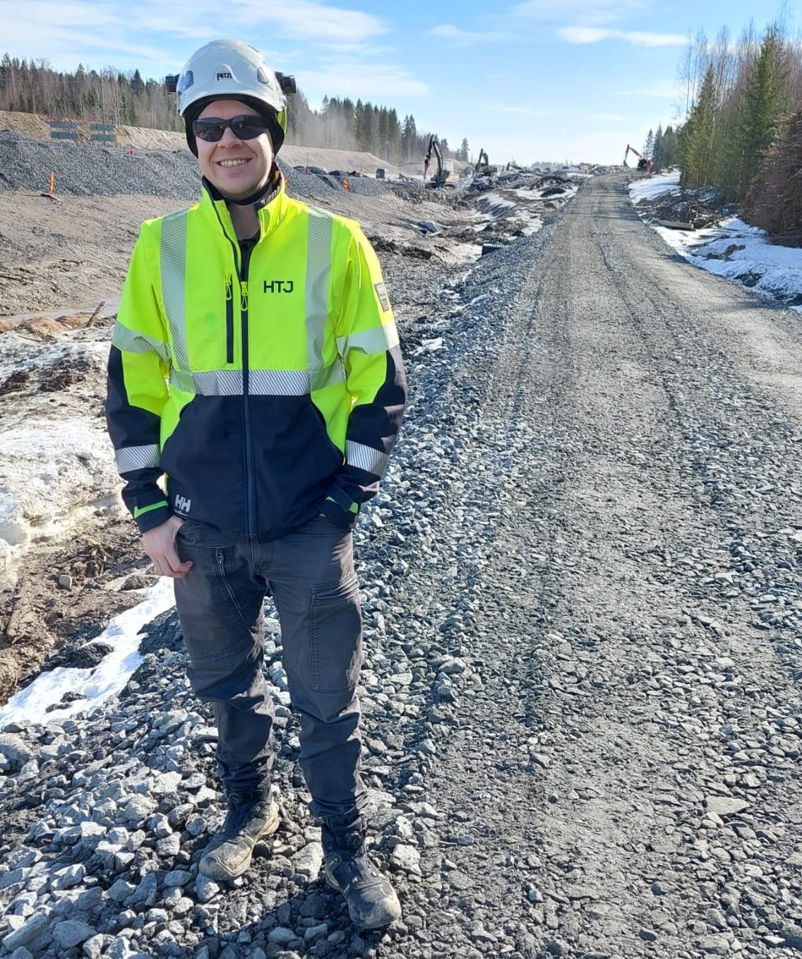 Rakennustyöntekijä seisoo hymyillen sorapintaisella tietyömaalla kirkkaassa kevätauringossa. Hänellä on yllään keltainen heijastava työtakki, työmaakypärä ja aurinkolasit. Taustalla näkyy rakennustyömaa, kaivinkoneita, lumisia penkkoja ja metsän reunaa.