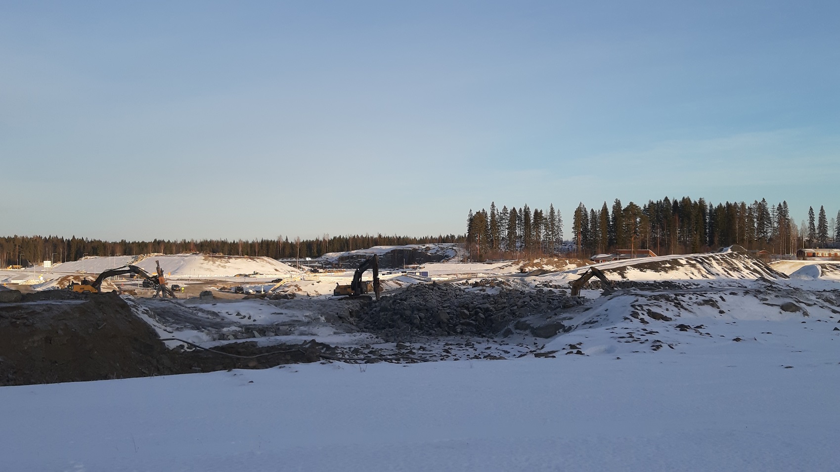 Kuvassa on luminen maisema, jossa kaivinkoneet työskentelevät avolouhoksessa, ja taustalla näkyy metsää.