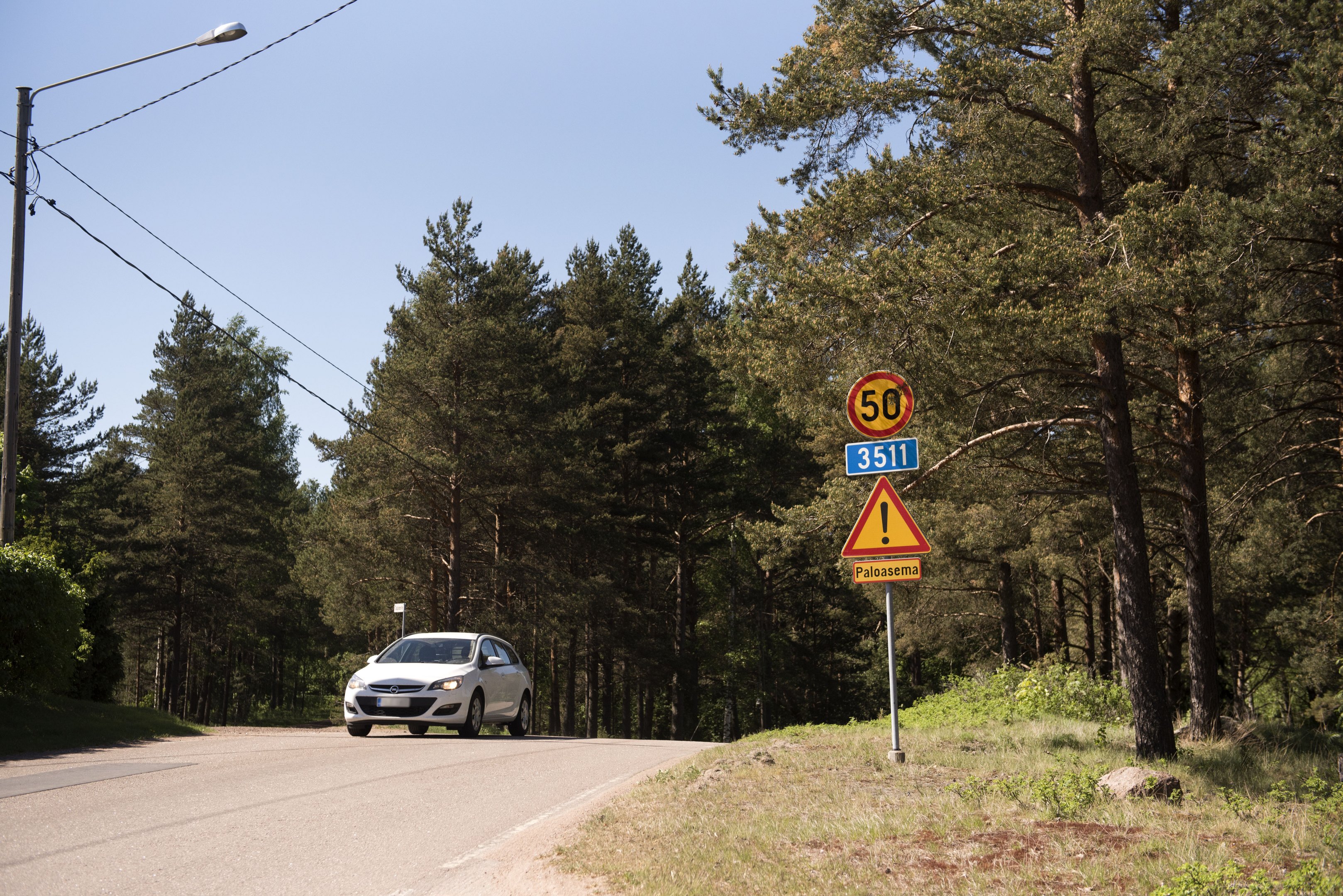 Mäntyjen ympäröimä tie, jolla on nopeusrajoitus 50 km/h. Tiellä ajaa valkoinen henkilöauto.