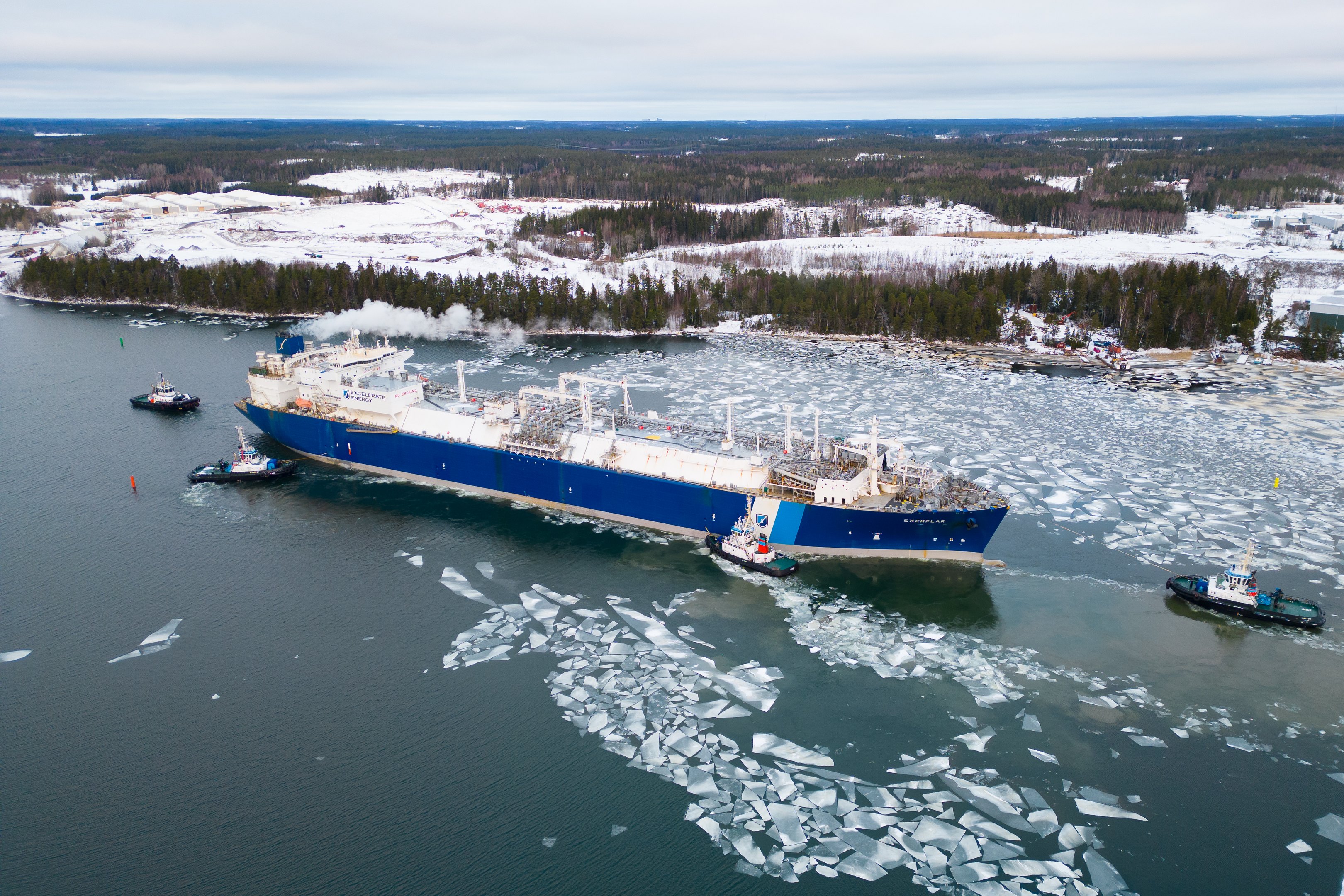 Nestetytettyä maakaasua kuljettava sinivalkoinen Exemplar-laiva lipuu eteenpäin jäätyneissä vesissä avustavien veneiden saattamana. Taustalla näkyy talvista metsää.