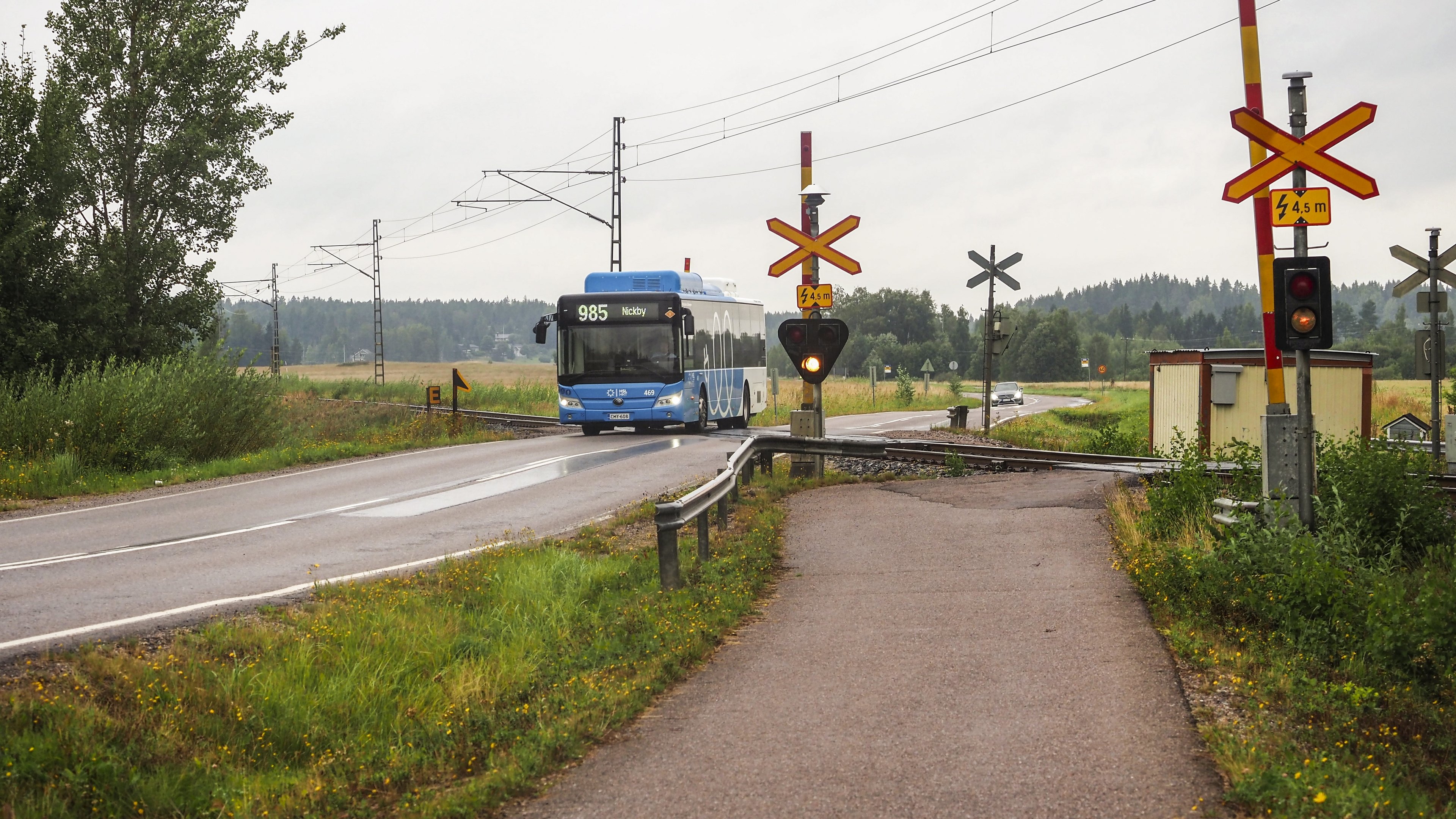 Bussi ylittää vartioitua tasoristeystä sateisessa säässä. Ympäristössä näkyy peltoa ja metsää.