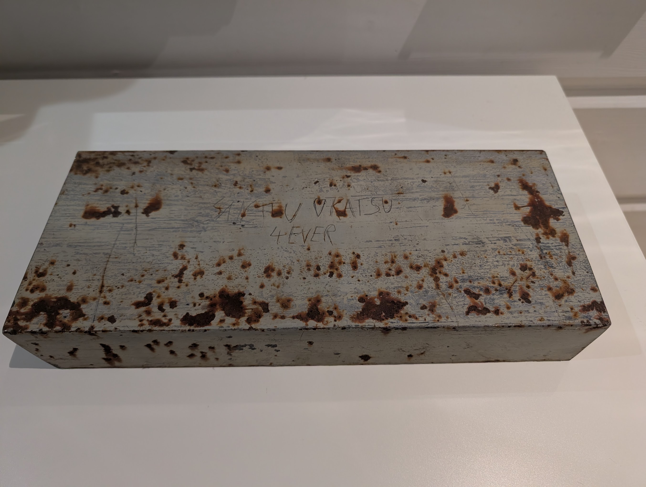 Ainonsolan sillan kaiteesta irrotettu metallipala sisätilassa. Pinnan ruosteessa näkyy käsin kaiverrettu teksti.