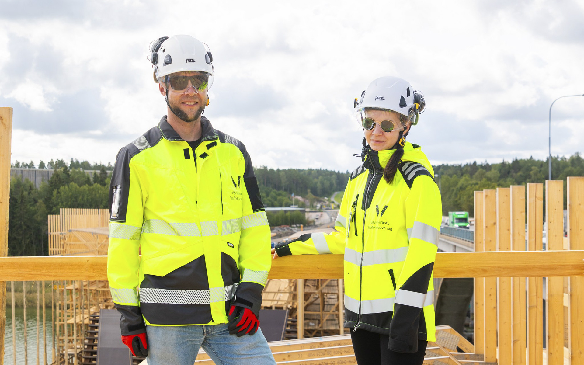 Kaksi henkilöä, joilla on yllään huomiotakit ja valkoiset kypärät, seisoo rakennustyömaalla. Taustalla näkyy puisia rakenteita ja osittain rakennettu silta sekä puita ja pilvinen taivas.