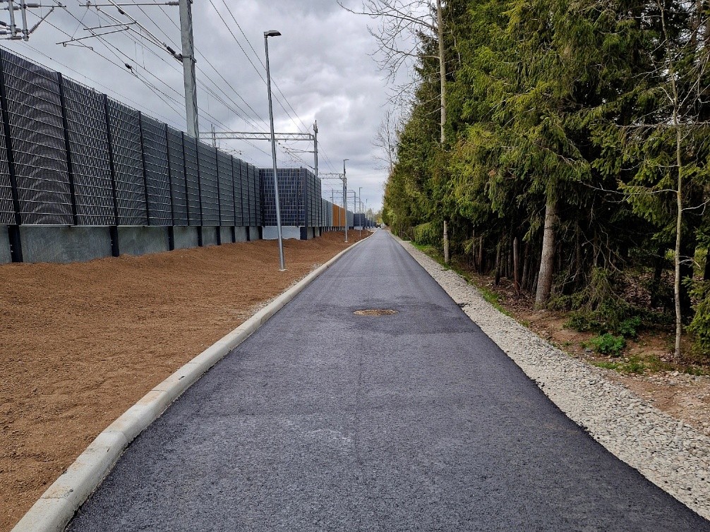 Asfaltoitu jalankulun- ja pyöräilynväylä, jonka vieressä maakaistale ja meluseinä. Toisella puolella väylää vihreää metsää.
