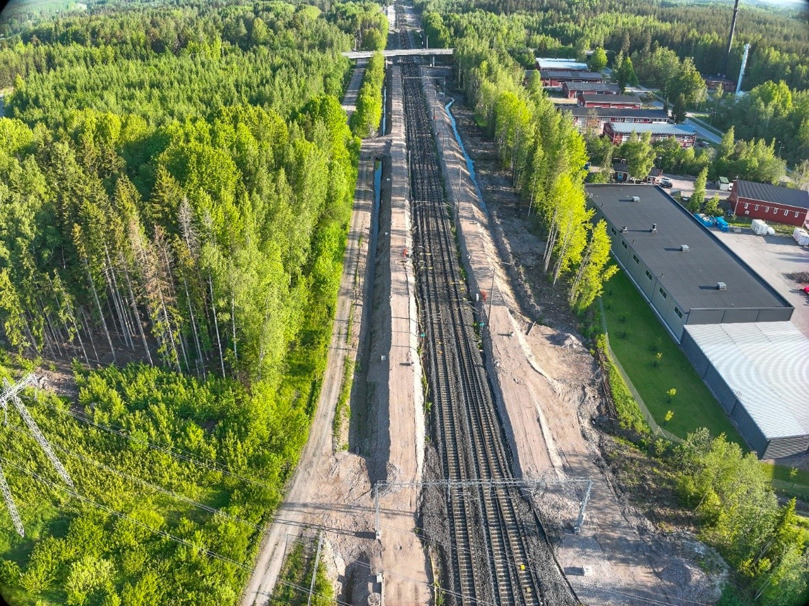 Esikuormituspenkereet ja nykyinen kaksoisraide kuvattuna ilmasta aurinkoisessa kesämaisemassa. Raiteiden vasemmalla puolella on kirkkaan vihreää metsää ja oikealla puolella teollisuushalli.