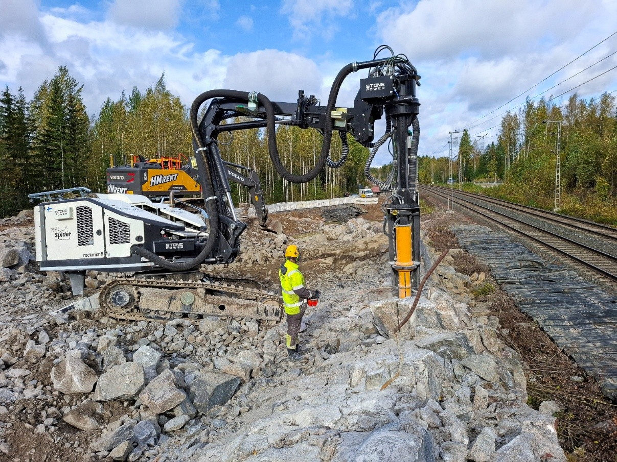 Kallio, jota porataan hydraulisella kiilalla puolipilvisessä säässä. Huomiovaatteisiin pukeutunut mies seuraa louhintaa kiilan vieressä. Kallion oikealla puolella kulkee kaksoisraide.