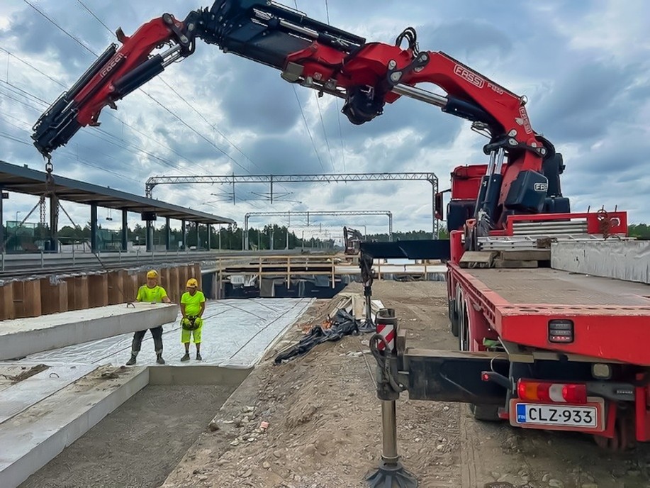 Nostoauto nostaa betonipalkkia sillan päälle. Kaksi keltaisiin huomiovaatteisiin pukeutunutta miestä seisoo laatan vieressä, toinen ohjaa laattaa oikealle paikalle. 
