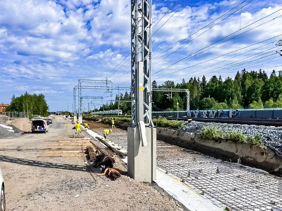 Radalla kulkee juna. Radan etupuolella keltaiseen huomiovaatteisiin pukeutuneet miehet tekevät paalulaatan raudoituksia aurinkoisena kesäpäivänä. Etualla näkyy kasassa raudoitteita.