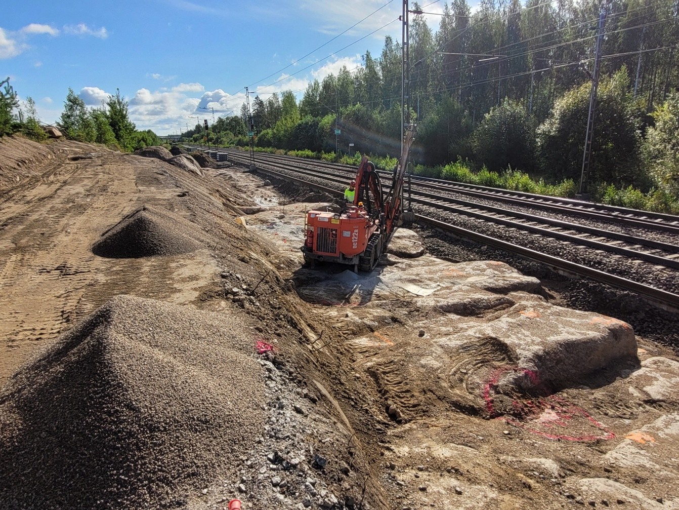 Raiteiden vieressä on louhittua kalliopintaa, ja siinä seisoo punainen työkone työntekijän kanssa jatkamassa louhintaa aurinkoisessa kesäsäässä.