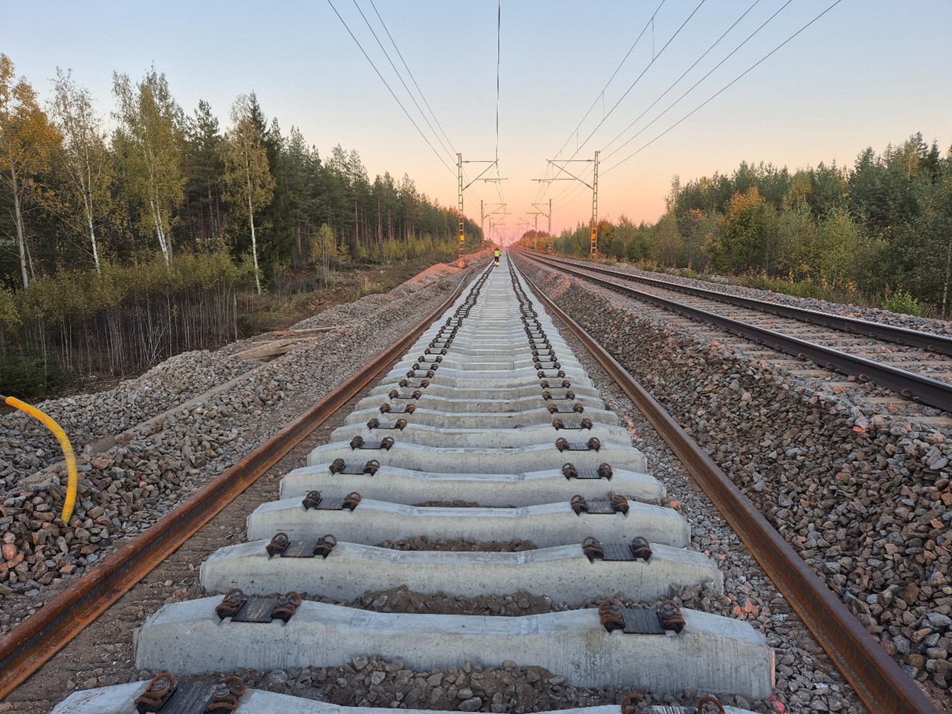 Taivas hehkuu pinkin sävyissä pilvettömänä kesäyönä. Kaksi raidetta kulkee vierekkäin. Toiselta niistä on poistettu vanha päällysrakenne ja asennettu osittain tilalle uudet pölkyt.