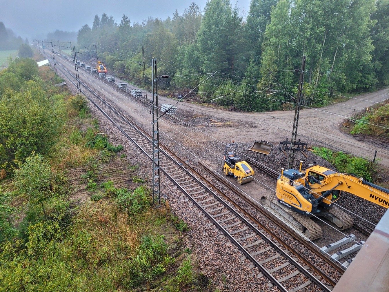 Kolme ratatyökonetta työskentelee raiteella ja sen vieressä syksyisessä ja sumuisessa ilmassa. Osa puista ja pensaista on alkanut jo kellertää.