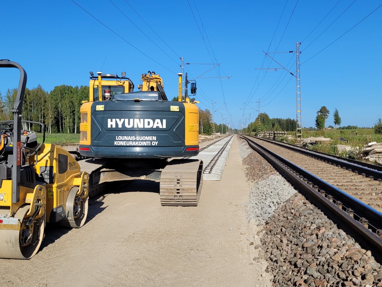 Vehreän peltomaiseman keskellä kulkee kaksi raidetta. Toisella raiteella seisoo kaksi keltaista työkonetta ja käynnissä on pölkkyjen asennus.