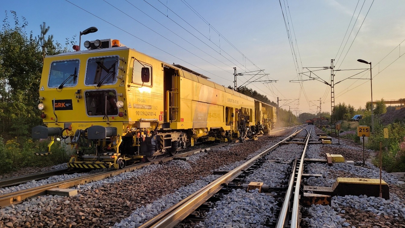 Ilta-aurinko paistaa matalalta elokuun illassa. Radalla seisoo keltainen tukemiskone.