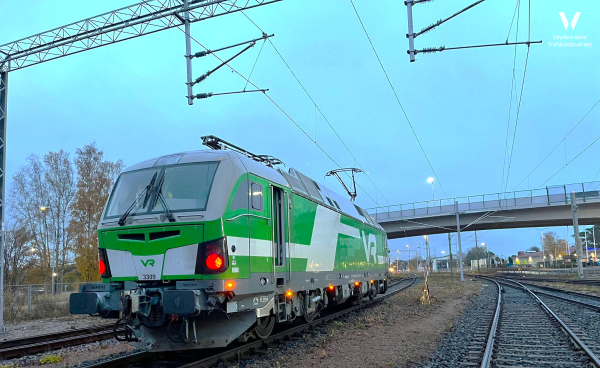 Kuvassa etualalla VR:n veturi, joka on juuri tullut ylikulkusillan alta. Rata on sähköistetty. Päivä on juuri menossa mailleen, mutta vielä on valoisaa.