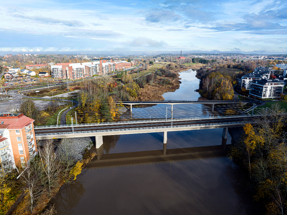 Ilmakuva joesta, jonka yli kulkee rautatiesilta ja sen yläpuolella kevyen liikenteen silta. Joen molemmin puolin näkyy syksyinen kaupunkimaisema, kerrostaloja, puita ja rautatiekiskoja, taustalla laaja kaupunkialue ja pilvinen taivas.
