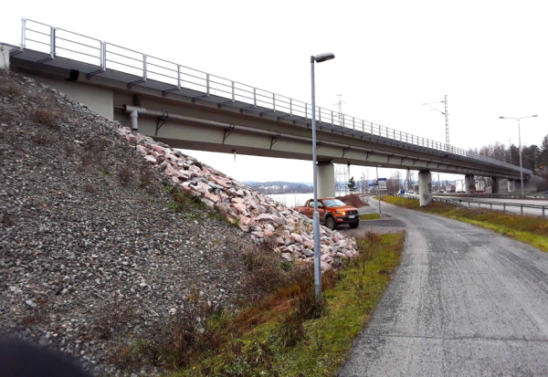 Harmaa, sateinen päivä, silta kulkee kuvassa vasemmalta oikealle, sillan alla on asfaltti- ja hiekkatie sekä punainen auto.