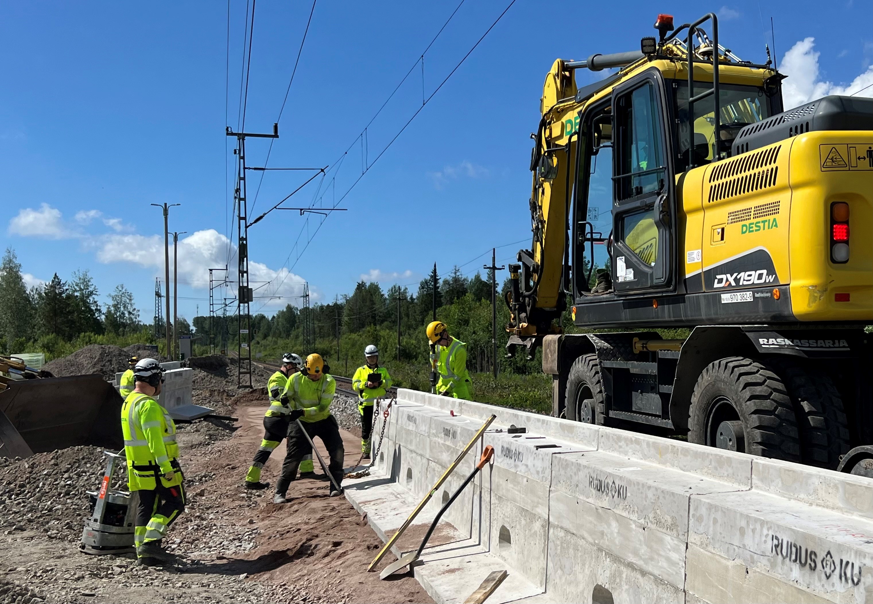 Rakennustyömaalla keltainen kaivinkone ja viisi huomioliiveissä olevaa työntekijää asentavat betonisia laiturielementtejä rautatien viereen. Taustalla näkyy radan sähköjohdot ja vihreää metsää.