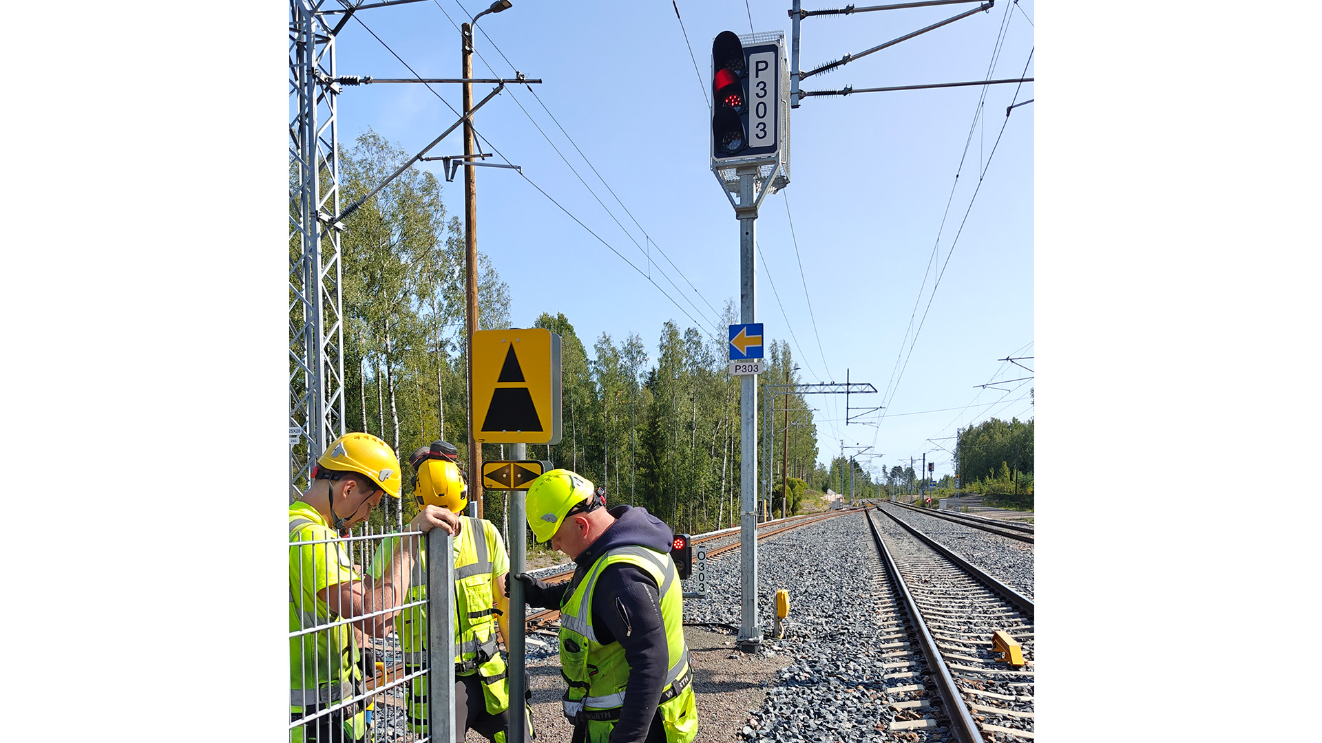 Kolme kypärään ja huomioliiviin pukeutunutta työntekijää tarkastelee asennettua ratamerkkiä. Taustalla näkyy rautatie, opasteita ja punaista näyttävä liikennevalo. Oikealla näkyy kaukaisuuteen jatkuva sähkörata. 