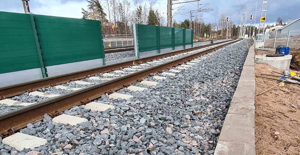 Kuvassa on junaraide ja vihreää meluaitaa. Radalla työskentelee työmiehiä.