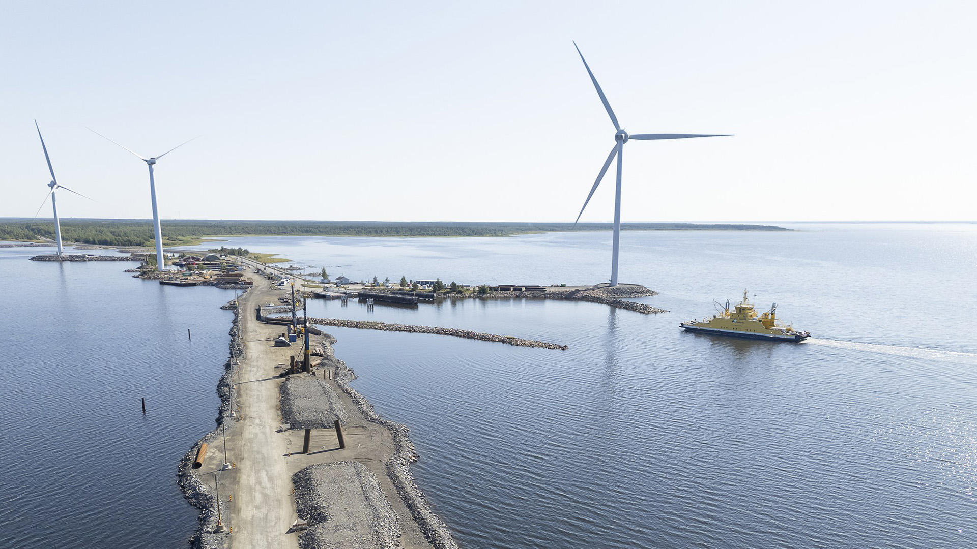 A quarry embankment under construction from the mainland to the sea, a ferry port next to it and a connecting ship on its way there, wind turbines in the background.