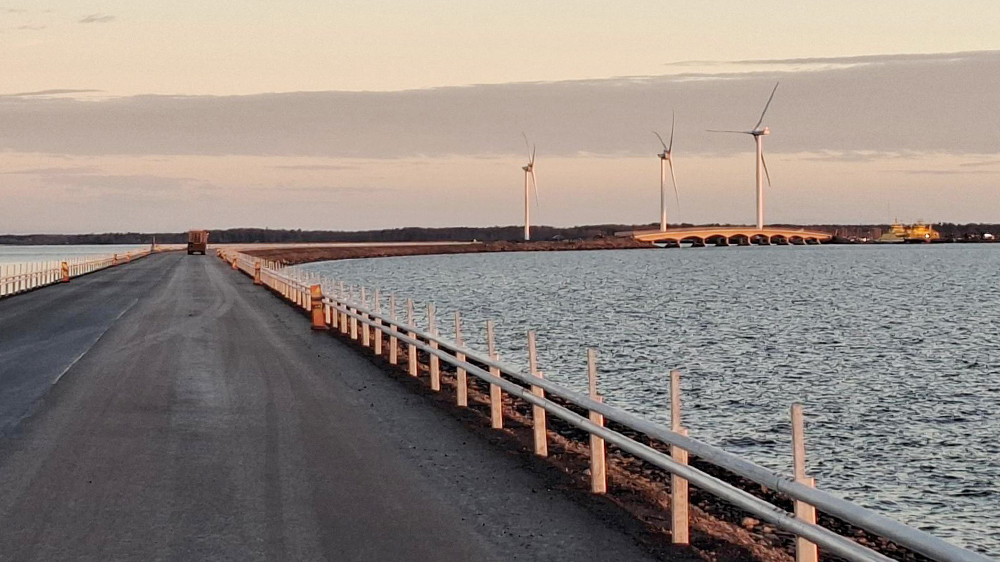 Kuorma-auto ajaa pengertiellä uudella asfaltilla kohti siltaa. Taustalla tuulimyllyjä ja lautta rannassa.