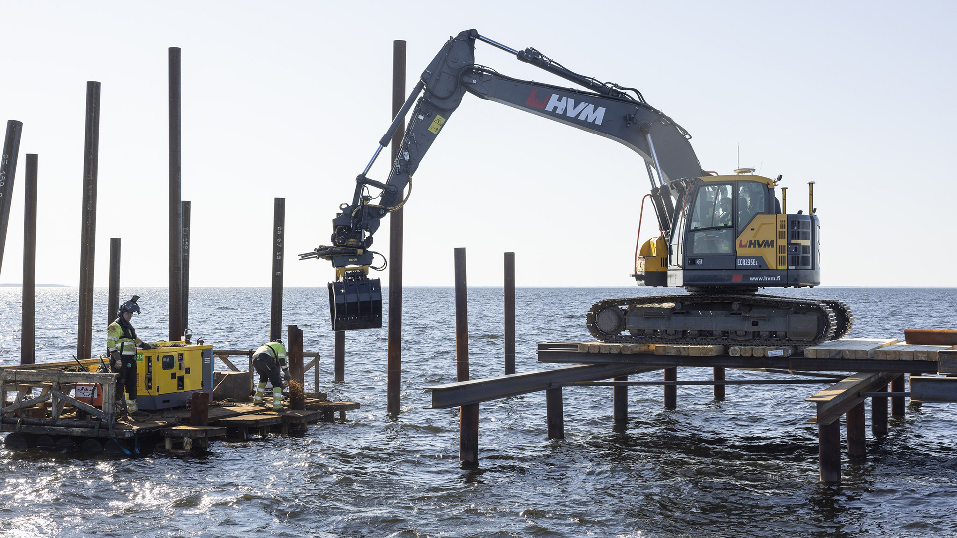Rakennustöitä merellä. Kaksi työntekijää työlautalla ja kaivinkone paalujen varassa lepäävien ponttilautojen päällä.