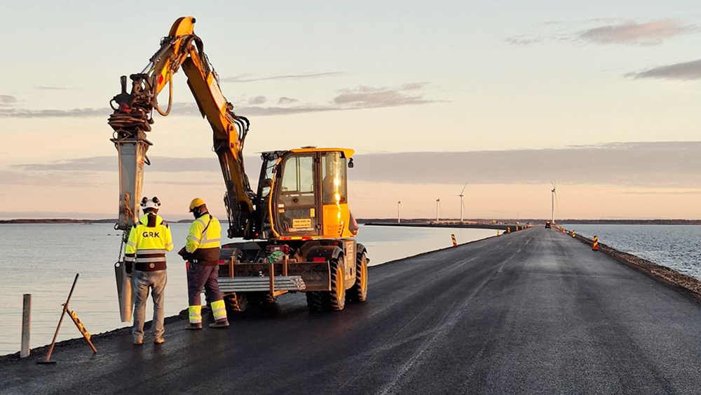 Työkone laittaa pengertien kaiteiden tolppia paikalleen uudella asfaltilla. Koneen vieressä seisoo kaksi henkilöä työvaatteissa. Taustalla merta ja tuulimyllyjä.