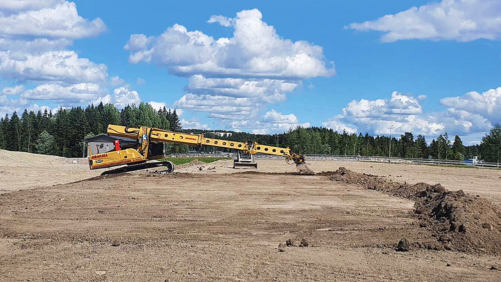 Keltainen kaivinkone, jolla on poikkeuksellisen pitkä puoli aukealla kentällä. Taustalla näkyy puita ja sinisellä taivaalla on poutapilviä.