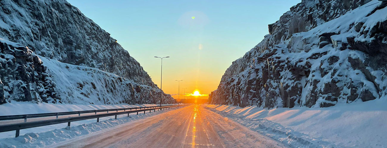 Tie halkoo kallioleikkausta, maassa on lunta, oranssina hehkuva aurinko on matalalla horisontissa.