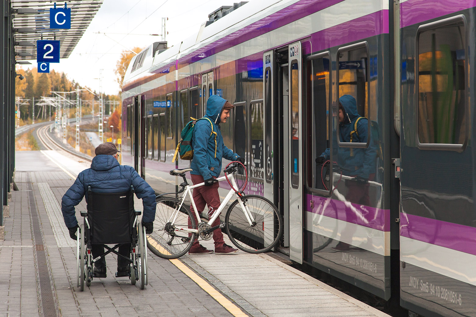 Kuvituskuva rautatieasemasta, jossa junaan astumassa pyörätuolinkäyttäjä ja pyöräilijä.